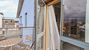 Un balcón con una mesa y sillas de madera, una puerta de cristal con cortinas y vistas a edificios cercanos.