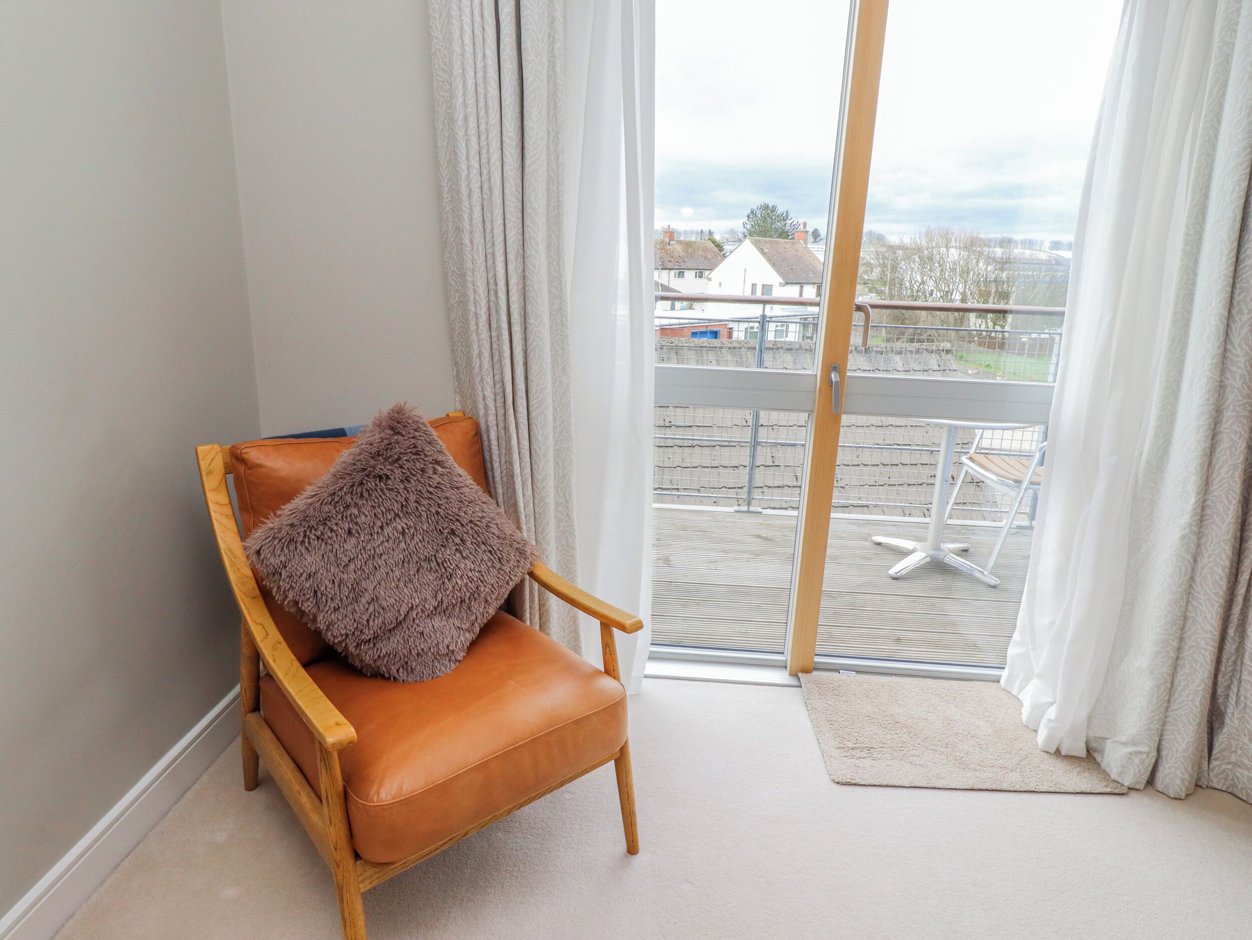 A room with a brown leather chair, a fluffy pillow, and a view of a balcony with a railing and a few houses in the background.
