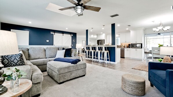Een moderne woonkamer met een grijze bank, een poef en een blauwe fauteuil, een keuken met witte kasten, en een plafondventilator.