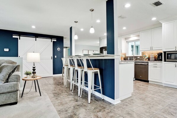 A modern kitchen with white cabinets, a central island with bar stools, and a dining area with a sofa and lamp.