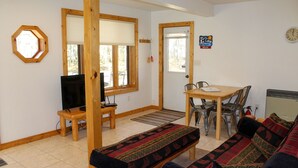 Een woonkamer met een bank, een televisie, een eettafel met stoelen en een raam met uitzicht op bomen.