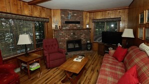 Un salón acogedor con una chimenea de ladrillo, paredes de madera, un sillón rojo, un sofá de cuadros y un televisor.