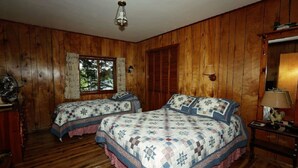 Una habitación con paredes de madera y dos camas con colchas. Hay una ventana con cortinas, una lámpara en una mesita de noche y una alfombra roja en el suelo.
