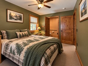 A bedroom with a bed, a ceiling fan, a window with blinds, a bedside table with a lamp, and a door.