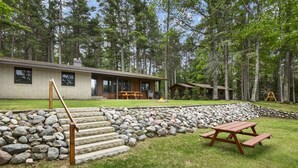 A cabin with a stone staircase leading to a wooden deck, surrounded by a stone wall and a grassy area with picnic tables.