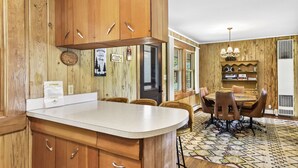 A kitchen with wooden cabinets, a white countertop, and a dining area with wicker chairs and a patterned rug.