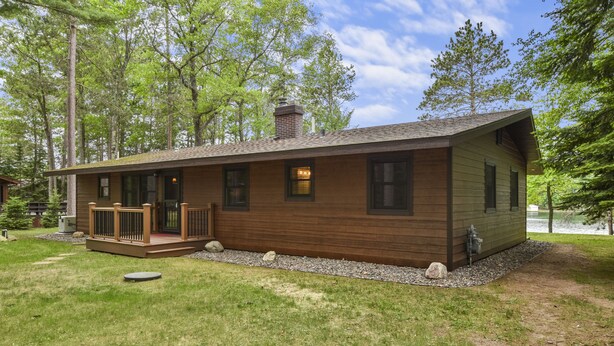 A wooden cabin with a deck, surrounded by trees and a grassy yard.