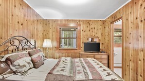 A wooden-paneled bedroom with a bed, a nightstand, a television, and a window with blinds.
