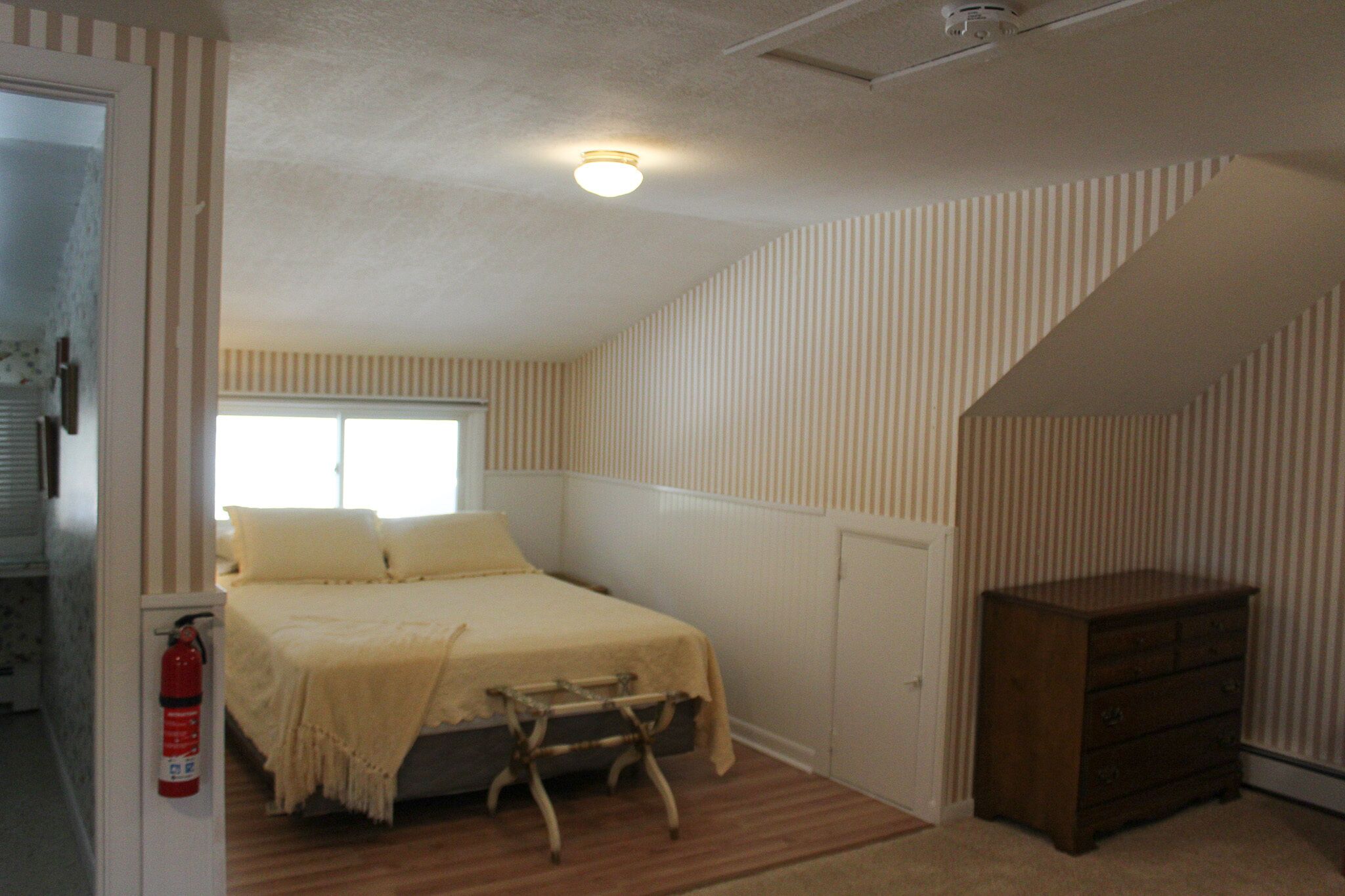 A bedroom with a bed, a wooden dresser, a window, and a fire extinguisher.