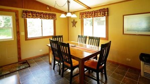 A dining room with a wooden table and chairs, large windows with curtains, a star decoration on the wall, and a painting. - Liam's Landing located on Little St Germain Lake (St Germain)