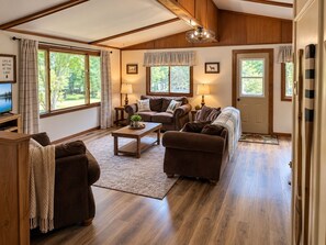Un salon chaleureux avec une cheminée, un plancher en bois et de grandes fenêtres donnant sur l’extérieur.