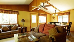 A living room with a brown sofa, a glass coffee table, a ceiling fan, and a dining area with a table and chairs. - Liam's Landing located on Little St Germain Lake (St Germain)