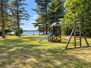 Un terrain de jeux avec un toboggan et des balançoires, entouré d’arbres et offrant une vue sur un lac.