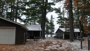 Un ensemble de chalets reliés par un chemin de terre menant à un garage, le tout entouré de grands arbres et avec un lac en arrière-plan.