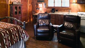 A rustic cabin interior with a bed, two leather chairs, a dining table, a kitchen area, and wooden walls. - Deer Run Unit #8 on Big St Germain Lake (St Germain)