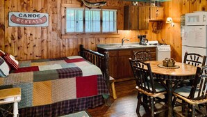 A cabin interior with a wooden floor, a bed with a quilt, a dining table with chairs, a kitchen with wooden cabinets, and a sign for canoe rentals. - Deer Run Unit #9 on Big St Germain Lake (St Germain)