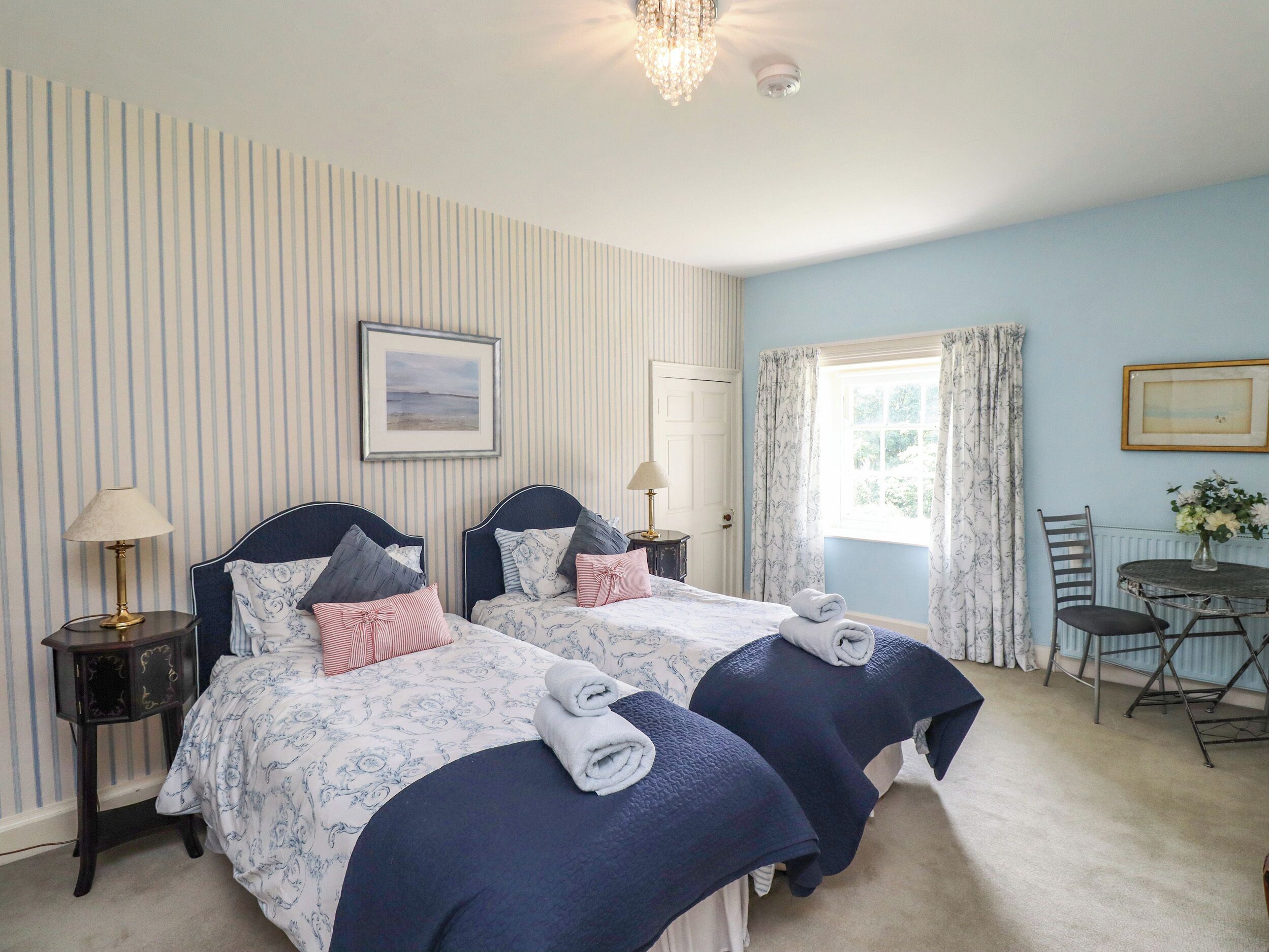A double room with two beds, a chandelier, a framed picture on the wall, and a window with curtains.
