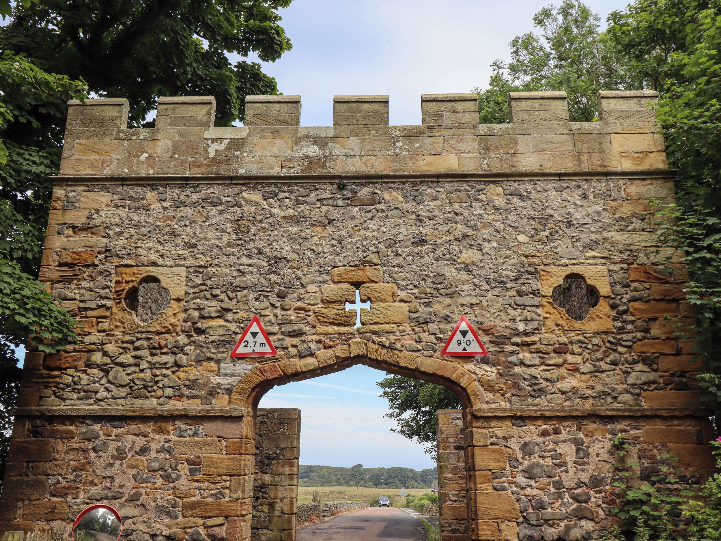 A stone archway with a cross symbol, flanked by triangular warning signs indicating a height restriction of 2.7 meters and a drop of 9-10 meters.