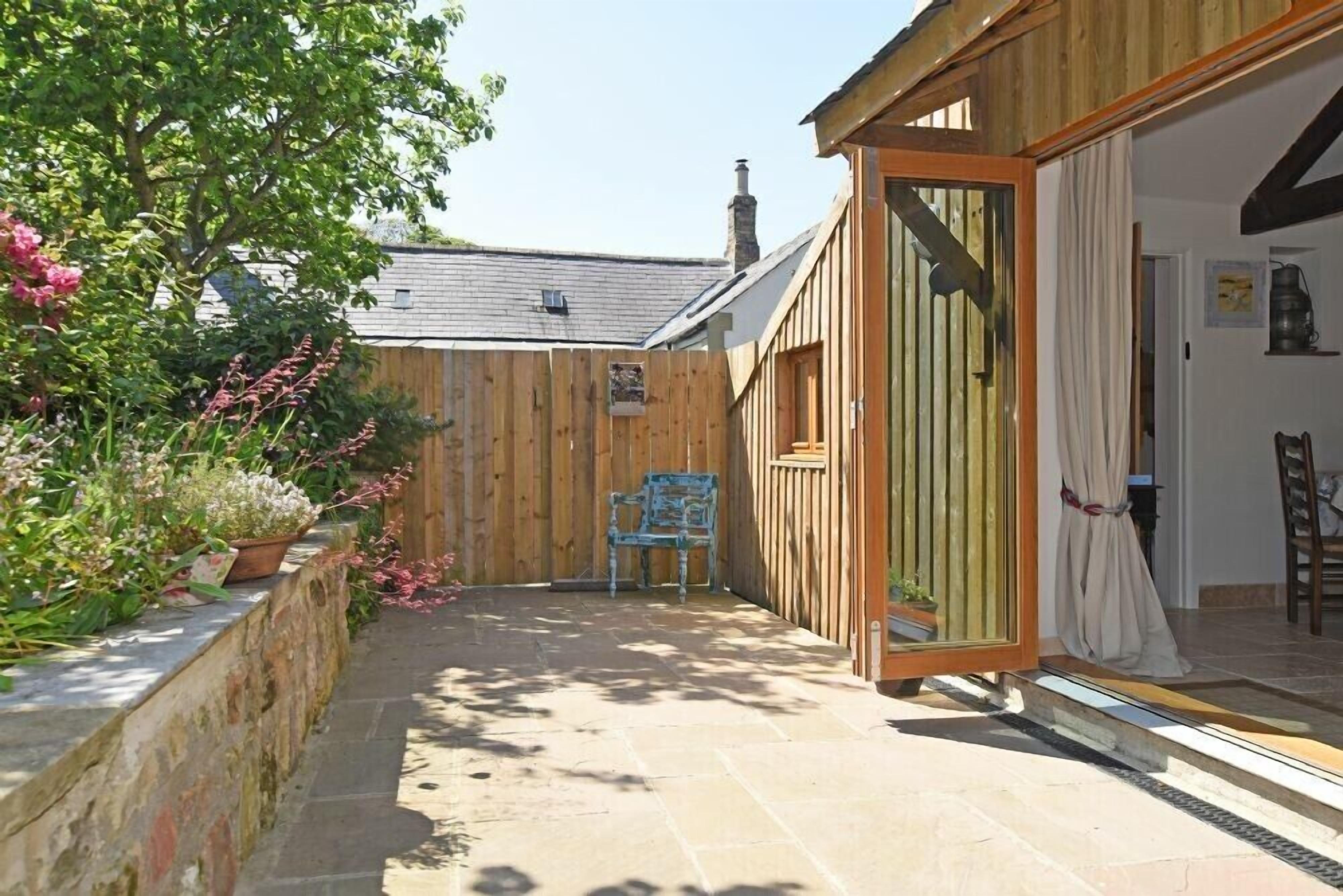 A wooden cottage with a stone patio, a wooden fence, and a garden with pink flowers.