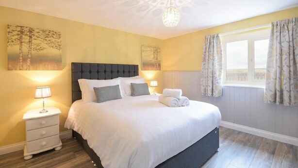 A neatly made bed with white linens, a dark headboard, and two bedside lamps. There is a window with curtains, a nightstand with a lamp, and artwork on the walls. - Dairymaid's Cottage (Morpeth)