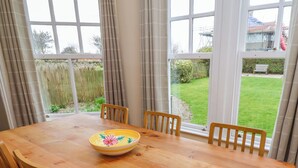 Une table à manger avec des chaises et un saladier, de grandes fenêtres avec des rideaux, et une vue sur une cour verdoyante.