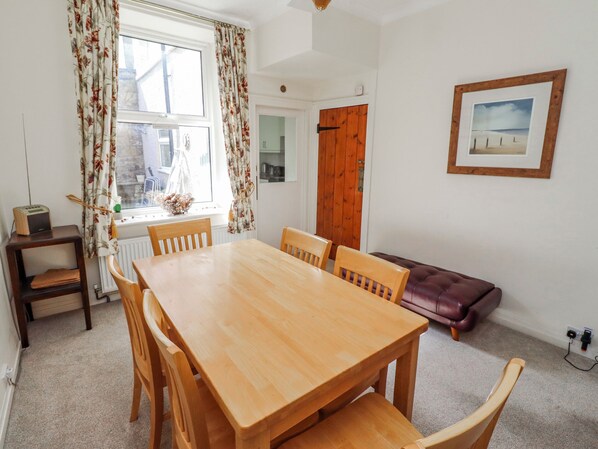 A dining area with a wooden table and chairs, a radiator, a window with curtains, a door, and a framed picture on the wall.