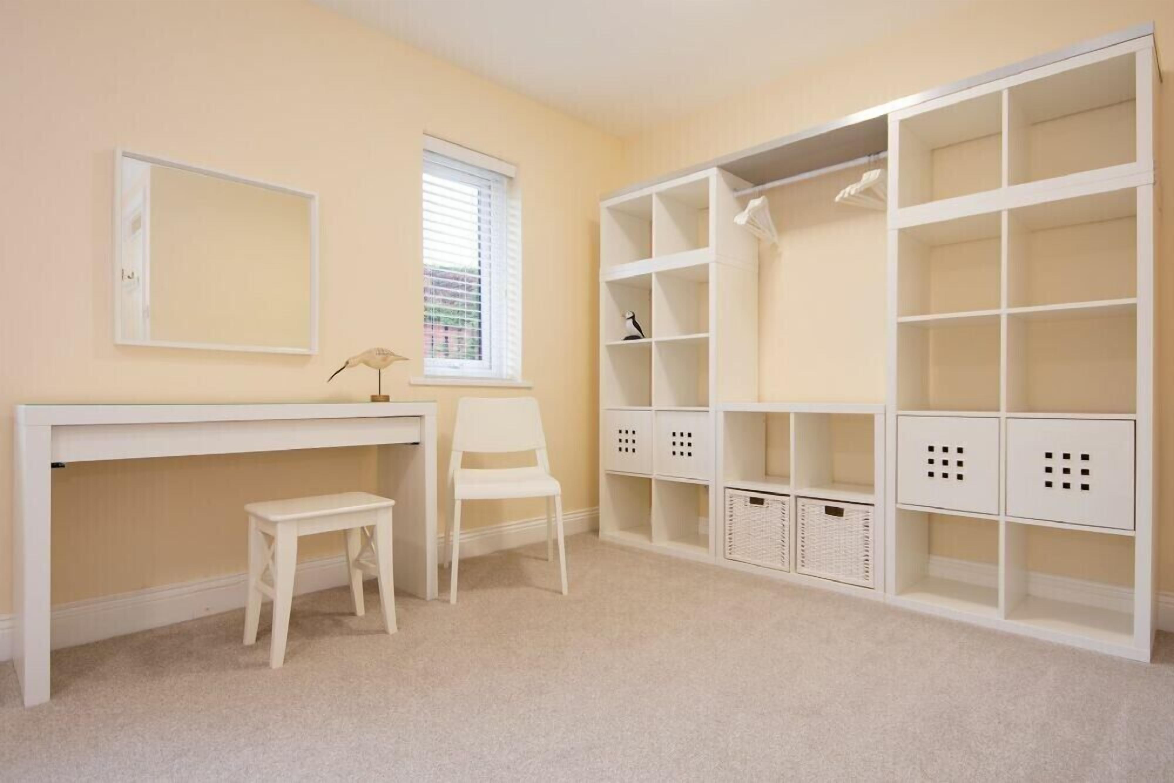 A room with a white desk, a chair, a small stool, and a large white shelving unit with baskets.