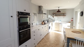 A modern kitchen with white cabinetry, a built-in oven, a washing machine, and a dining area with a wooden table and chairs. - The Hollies (Bamburgh)
