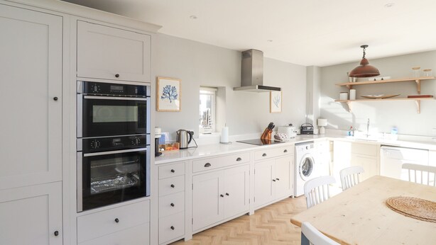 A modern kitchen with white cabinetry, a built-in oven, a dishwasher, and a wooden dining table. - The Hollies (Bamburgh)