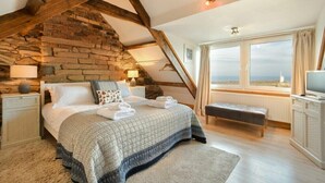 A bedroom with a stone wall, wooden beams, a large bed, a bench, a TV, and a window with a view. - Farne Lookout (Seahouses)