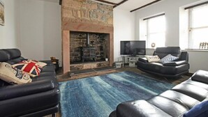 A living room with a stone fireplace, leather sofas, a television, and a large window. - Beach Retreat (Seahouses)