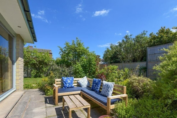 Une terrasse avec un banc en bois et une table, entourée d’une végétation luxuriante et d’une clôture en bois.