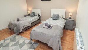 A bedroom with two beds, each with a nightstand, a lamp, and a pillow. There is a painting on the wall and a patterned rug on the floor. - Butcher's Retreat (Seahouses)