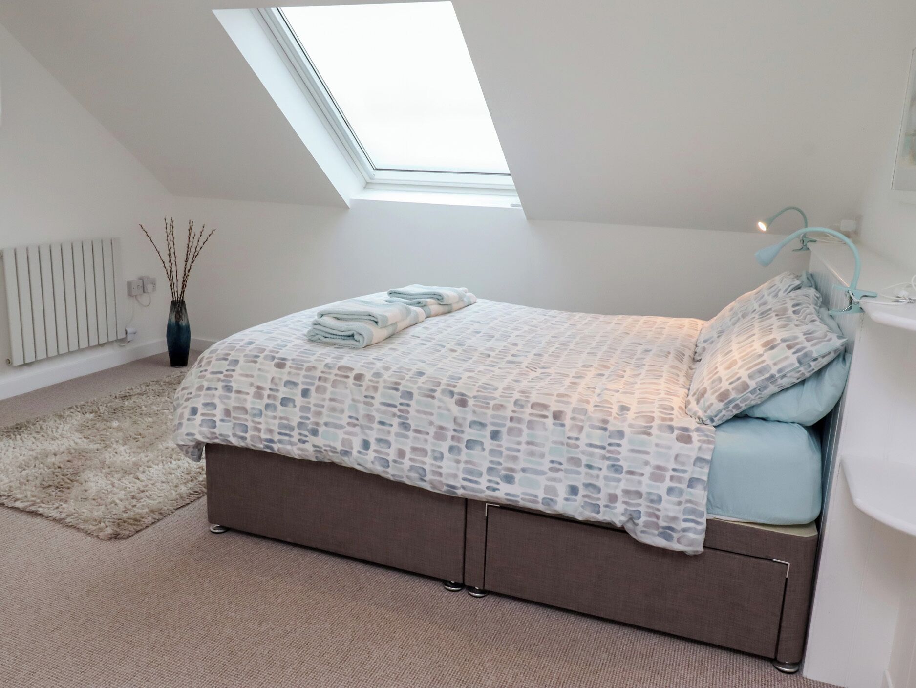 A bedroom with a skylight, a bed with a patterned bedspread, a bedside table, and a radiator.