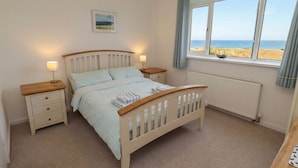A bedroom with a bed, bedside tables, a window with a view of the sea, and a radiator. - Dell Point (Chathill)