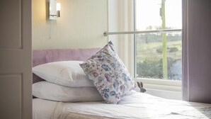 Ein Bett mit weißer Bettwäsche und einem blumigen Kopfkissen, eine Nachttischlampe und ein Fenster mit Blick auf ein Wiesenfeld.