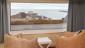 Une pièce dotée d’une grande fenêtre donnant sur une côte rocheuse et la mer. On y trouve deux fauteuils en osier et une petite table blanche.