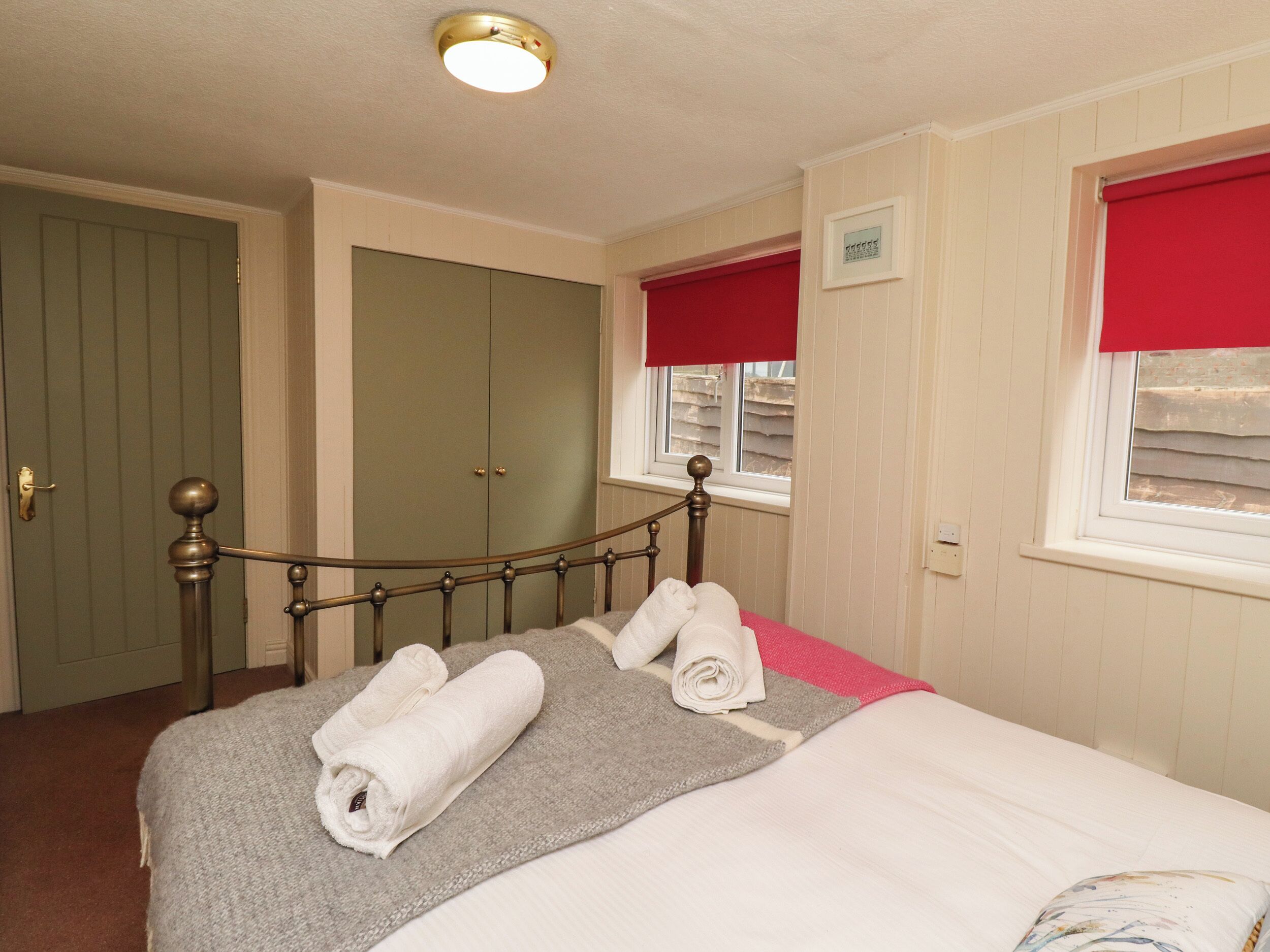 A neatly made bed with rolled towels, a window with red blinds, and a door with a brass handle.