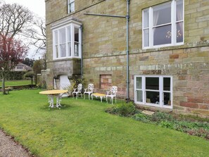 A stone building with a corner window and a bay window. There is a small garden with a table and chairs. - Glebe Cottage (Bamburgh)