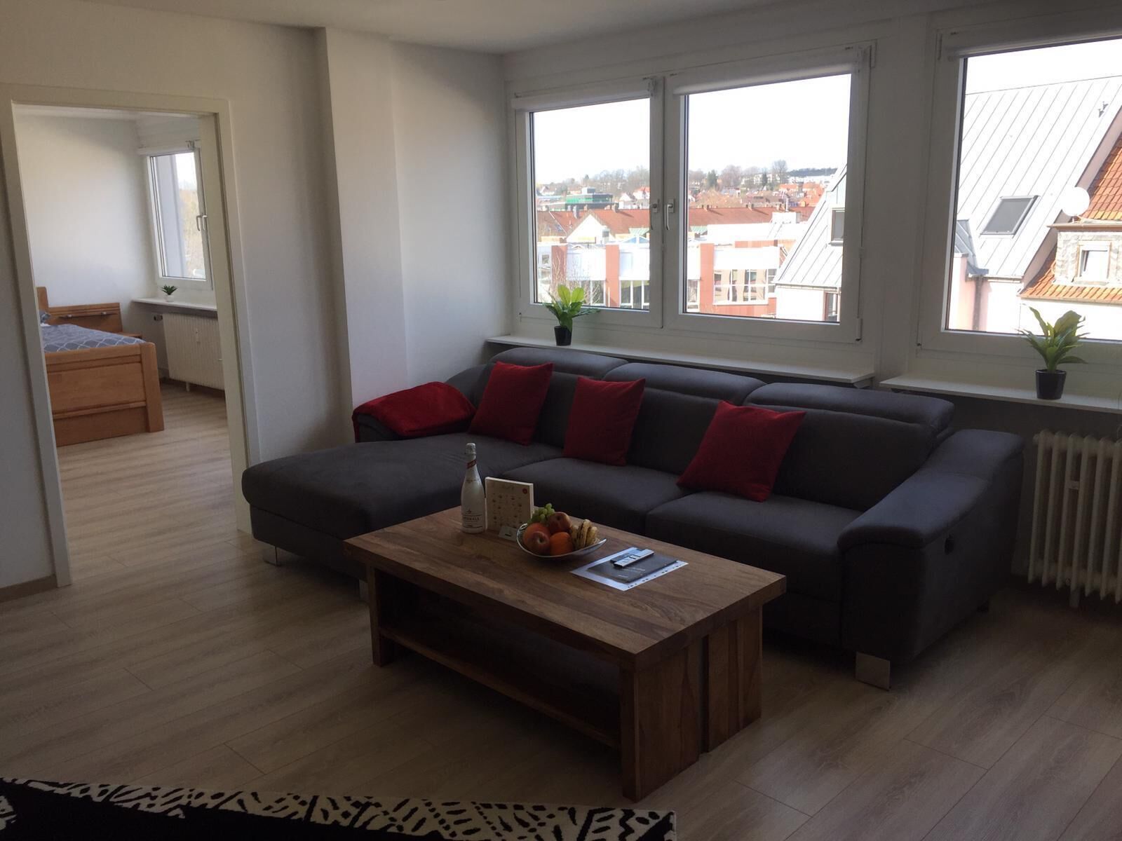 A modern living room with a grey sofa, a wooden coffee table, and a view of buildings through large windows.