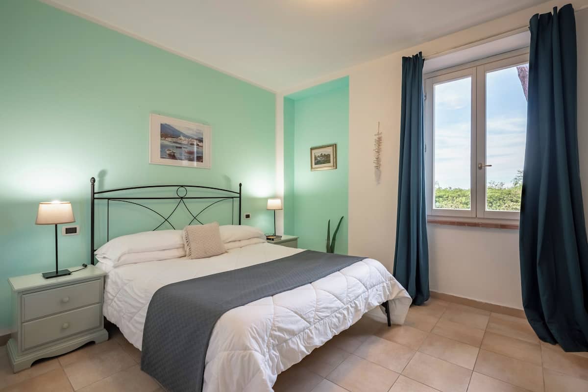 Un dormitori amb llit, fallegueres, una finestra amb cortines i dos quadres penjats a la paret.
