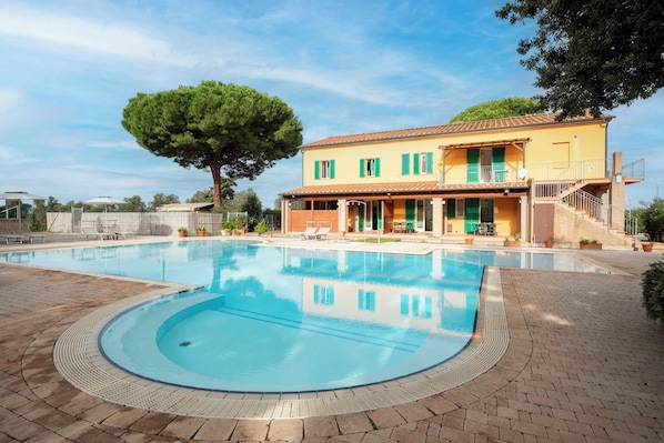 A swimming pool with a tiled deck, a two-story building with green shutters, and a large tree in the background.