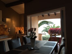 Ein Essbereich mit Tisch für vier Personen, einer Blumenvase und Blick auf einen Garten mit Palmen und roten Blättern.