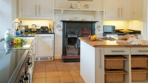 A kitchen with white cabinets, a black stove, a tiled floor, a fireplace, and a countertop with a kettle and a vase of flowers. - The Villas No3 (Alnwick)