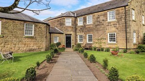 A stone building with a central entrance, a paved pathway, and a small garden area with benches and potted plants. - Treetops, Alnwick (Alnwick)