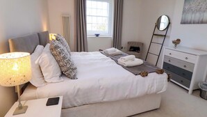 A neatly made bed with pillows, a bedside lamp, and towels. There is a window with curtains, a radiator, and a dresser with a mirror. - Treetops, Alnwick (Alnwick)