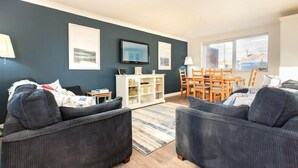 A living room with dark blue walls, a white entertainment unit, a dining table with chairs, and a large window.