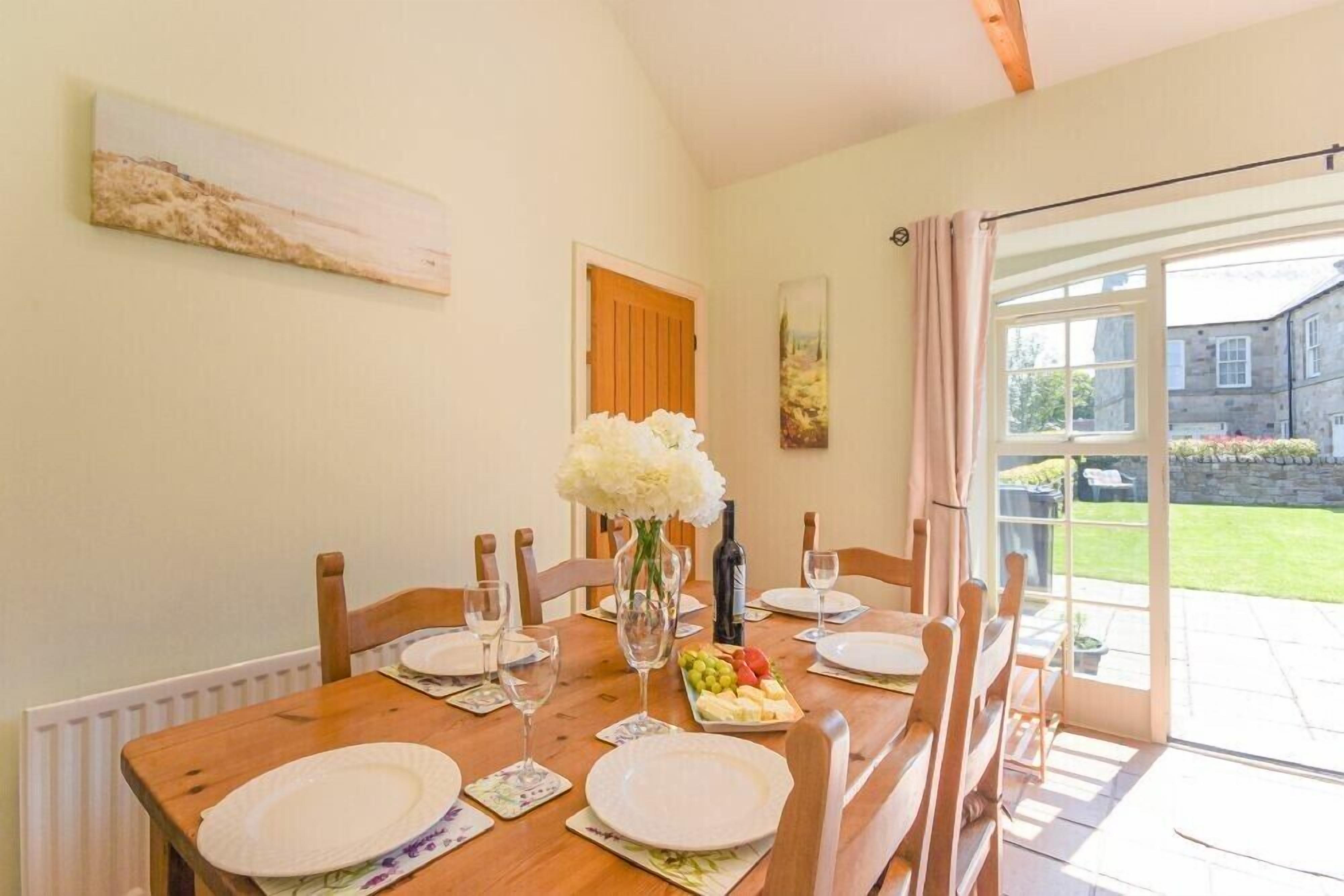 A dining area with a wooden table set for four, a vase with white flowers, and a view of the garden through the door.