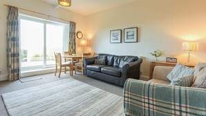 A living room with a sofa, a plaid armchair, a dining table with chairs, and a large window with curtains.