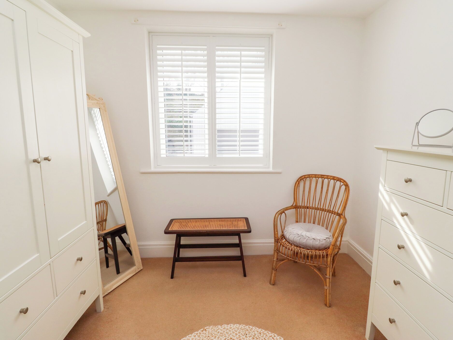 A room with a white wardrobe, a wicker chair, a small bench, and a dresser with drawers.